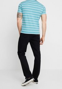 Camisa de rayas azules y blancas de manga corta combinada con pantalones negros y zapatillas a rayas, con un tatuaje en el brazo izquierdo.