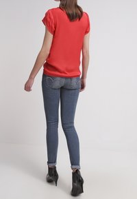 Frau trägt eine rote kurzärmelige Bluse, schmale blaue Jeans mit Herzstickerei auf den hinteren Taschen und schwarze Absatz-Stiefeletten, steht mit dem Rücken zur Kamera.