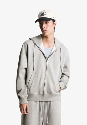 Svetlo siva zip-up kapuca z prostornim kapuco in sprednjimi žepi, izdelana iz mehke tkanine. V kombinaciji z sivo majico in bež bejzbol kapo.