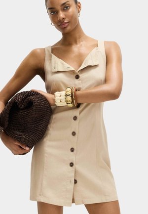 Frau trägt ärmelloses beiges Kleid mit Knopfleiste und hält große dunkle gewebte Tasche, dazu trägt sie breite Armbänder und goldene Creolen.