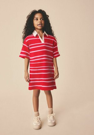 Nuori tyttö, jolla on pitkät kiharat hiukset ja joka pukeutuu puna-pinkkiraitaiseen mekkoon, beigen sävyisiin sukkiin ja valkoisiin tennareihin, seisoo yksivärisen taustan edessä.