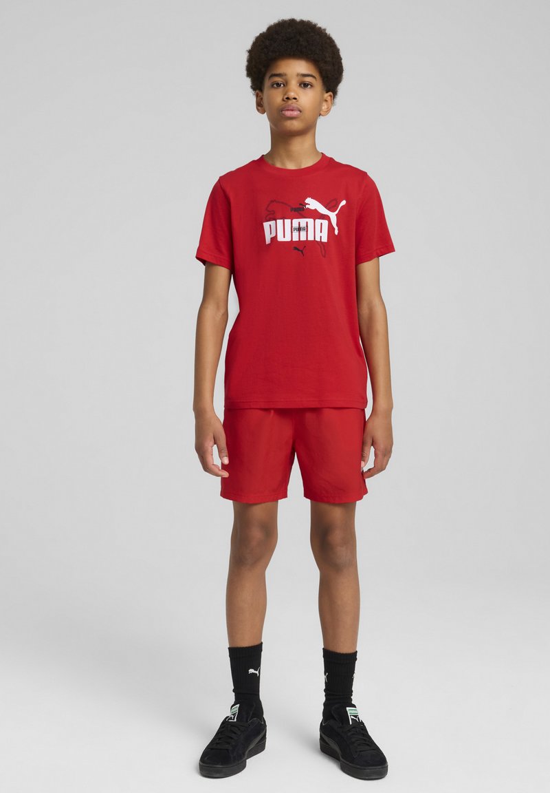 Camiseta de algodón roja y pantalones cortos con un logo de Puma en blanco. Llevado con zapatillas negras y calcetines. Diseño simple, mangas cortas y corte relajado.