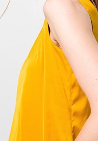 Seitenansicht einer Person in einem ärmellosen, leuchtend gelben Oberteil, das sich auf die Schulter und den Oberarm vor einem einfachen hellen Hintergrund konzentriert.