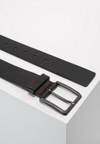 Curea din piele neagră cu o cataramă din metal mat, având accente de cusătură roșie și cinci orificii ajustabile pe lungimea sa.