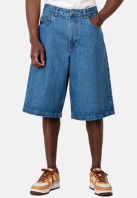 Pantalones cortos de mezclilla holgados, de color azul claro, hasta la rodilla, con un diseño clásico de cinco bolsillos y costuras visibles. Combinados con zapatillas que incluyen acentos marrones.