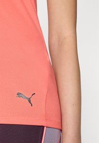 Korálové sportovní tričko s přiléhavým střihem, vyrobené z lehké tkaniny. Na dolním lemu se nachází černé logo Puma, spolu s fialovými legínami.
