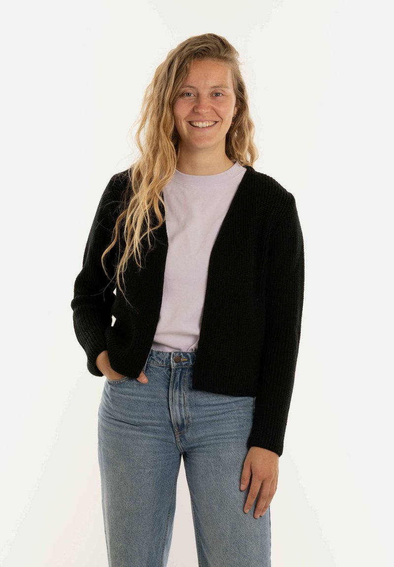 Zwarte gebreide cardigan met lange mouwen, open voorkant, gedragen over een lichtpaarse t-shirt en blauwe high-waisted jeans. Gladde textuur.