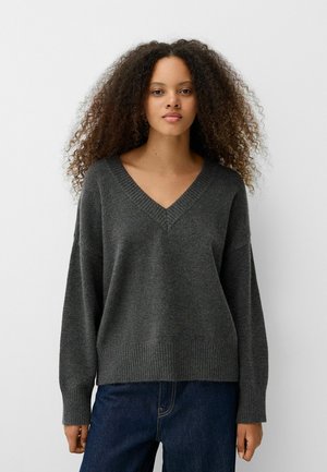 Gray, V-neck oversized sweater with dropped shoulders, ribbed cuffs, and hem. Soft texture with a relaxed fit, worn over dark jeans.