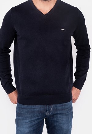 Mann trägt einen einfarbigen dunkelblauen V-Ausschnitt-Pullover mit kleinem Baum-Logo auf der Brust und blaue Jeans, Hände teilweise in den Taschen.