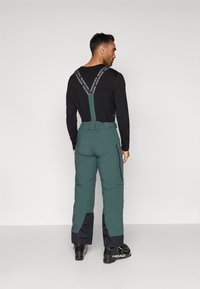 Calças de esqui verdes com suspensórios ajustáveis, apresentando detalhes pretos e um bolso com fecho de correr na lateral. Feitas de material durável e resistente às intempéries.