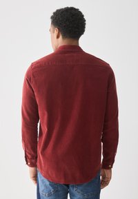 Camisa de pana roja con mangas largas, puños abotonados y cuello puntiagudo. Presenta un dobladillo curvado y una textura sutil.