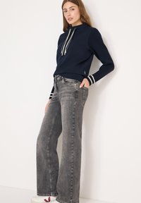 Marineblauer Pullover mit hohem Kragen und weißen Kordeln, kombiniert mit grauen ausgestellten Jeans und weißen Sneakers mit roten Akzenten.