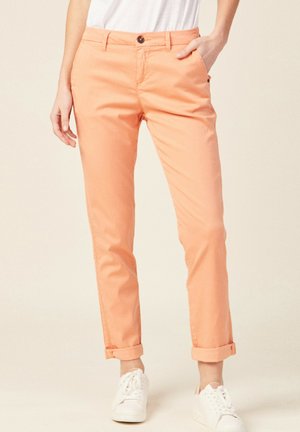 Persoon die opgerolde perzikkleurige broek, witte sneakers en een wit shirt draagt, staand met één hand in de zak.