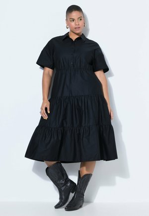 Žena v černých vrstvených midi šatech s krátkými rukávy a černých vzorovaných kovbojských botách stojící na bílém pozadí.