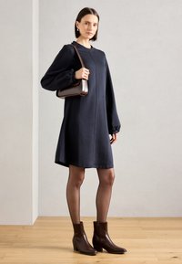 Marineblauwe jurk met lange mouwen, een losse pasvorm, textuurstof en knielengte; gecombineerd met een paar bruine enkelboots en een bruine schoudertas.