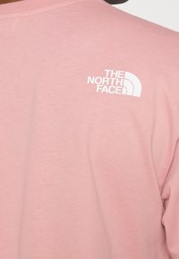 Lichtroze langarm t-shirt met een ronde hals en witte logotekst en ontwerp op de bovenrug. Zachte katoenstof met een gladde textuur.