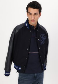 Veste universitaire marine avec manches en cuir, col côtelé avec des rayures bleues et blanches, boutons-pression et détail brodé sur la poitrine.