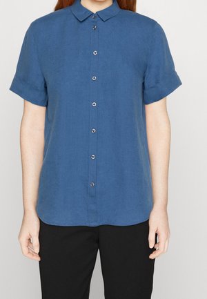Persoon draagt een blauw shirt met korte mouwen, knopen en kraag, en een zwarte broek, staand tegen een effen achtergrond.