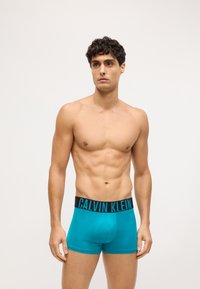 Boxeri de culoare teal cu un brâu negru pe care este inscripționat "CALVIN KLEIN" în albastru, confecționați dintr-un material neted și elastic, oferind o ajustare confortabilă.