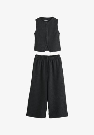 Ujjatlan fekete, texturált, gombos mellény párosítva hozzáillő bő szárú, elasztikus derekú nadrággal, fehér háttéren lefektetve.