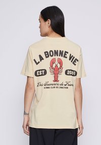 Beige T-skjorte med en rød hummergrafikk og svart tekst som sier "LA BONNE VIE EST 2001" på baksiden. Korte ermer, avslappet passform.