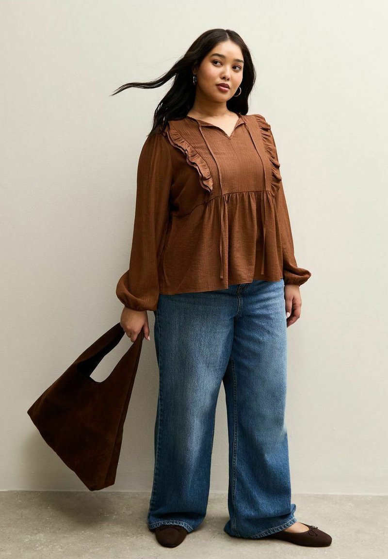 Brown, long-sleeved blouse with ruffled detail, paired with wide-leg blue jeans. Person holds a brown shoulder bag.