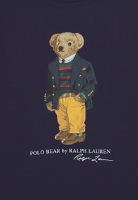 Teddy bear dressed in navy blazer, striped sweater, yellow pants, and brown shoes, standing with one hand in pocket on dark background.