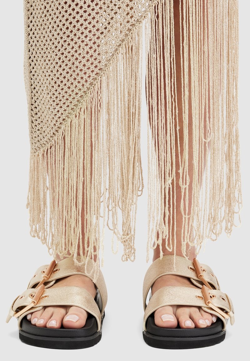 Pieds en sandales beige avec de grandes boucles, partiellement recouverts d'un tissu tricoté beige avec de longues franges pendantes.