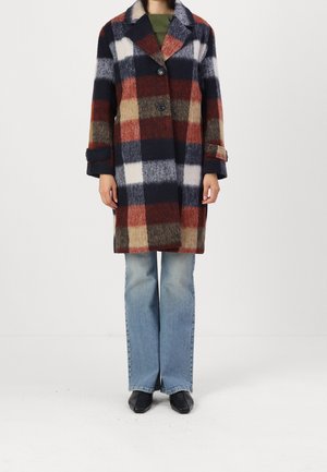 Kvinde iført en knælængde ternet frakke med store felter i marineblå, rust, creme og brun over falmede blå jeans og sorte spidse sko.