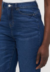 Blå denimjeans har høy midje, tettsittende silhuett og tradisjonell fem-lommers design med gule sømmer og sølvfarget metall.