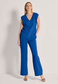 Blaues, kurzärmliges V-Neck-Oberteil, kombiniert mit passenden weiten Hosen, beide aus glattem, leichtem Stoff. Schwarze Sandalen mit Riemchen runden das Outfit ab.