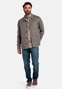 Veste à carreaux en tissu marron et beige, dotée d'une fermeture à bouton, d'une poche poitrine, et assortie à un jean bleu et des baskets grises.