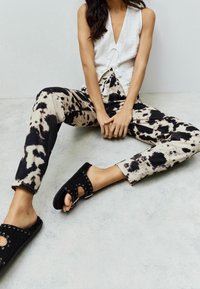 Haut décontracté à nouer devant en tissu blanc, associé à un pantalon beige et noir à effet tie-dye. Sandales plates noires avec découpes et accents métalliques.