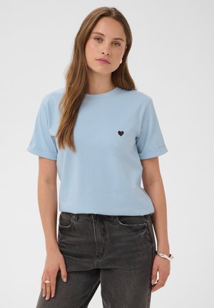 Kvinde iført en lyseblå T-shirt med et lille sort hjerte på brystet og mørkegrå jeans, stående mod en ensfarvet baggrund.