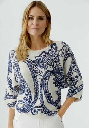 Frau mit blondem Haar, die einen weißen Strickpullover mit blauen, kunstvollen Paisley-Mustern trägt, die Hände in den Taschen, vor einem schlichten hellen Hintergrund.