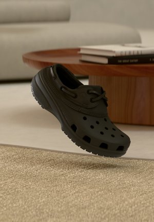 Μαύρο παπούτσι τύπου clog χωρίς κορδόνια με οπές αερισμού, τοποθετημένο πάνω σε υφασμένο μπεζ χαλί κοντά σε στρογγυλό ξύλινο τραπέζι με βιβλία.