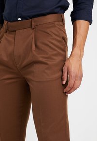 Bruna skräddarsydda byxor med glatt textur, med vecken, platt front och en slim fit-design. Matchas med en mörkblå skjorta.