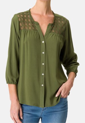 Olijfgroene blouse met doorschijnend geborduurd inzetstuk, knopen aan de voorkant, driekwart mouwen, gedragen met een blauwe spijkerbroek, één hand in de zak.