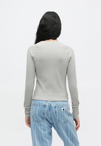 Γυναίκα με μακριά μαύρα μαλλιά, φορώντας ένα ανοιχτό γκρί ριγωτό μπλουζάκι με μακριά μανίκια και ανοιχτό μπλε ριγέ τζιν, γυρισμένη προς τα πίσω σε λευκή απλή φόντο.