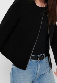 Veste noire à fermeture éclair sur le devant, avec une texture lisse, un col rond et un léger rembourrage aux épaules, portée avec un haut noir et un jean bleu.