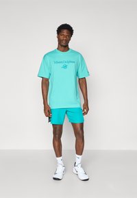 Camiseta turquesa con el texto y el logo de "Miami Dolphins", combinada con pantalones cortos a juego. El material parece suave; el diseño presenta un ajuste relajado.