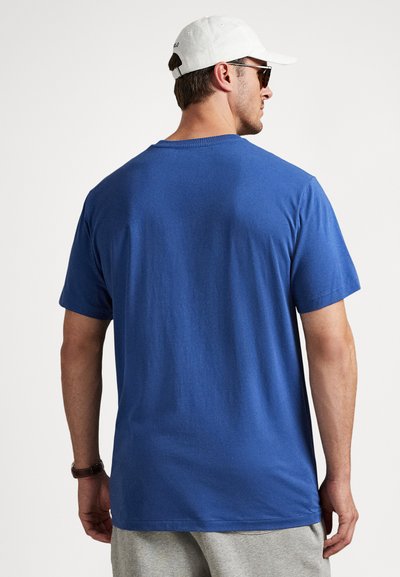 Bărbat purtând tricou albastru, pantaloni scurți gri, șapcă de baseball albă, ochelari de soare și ceas de mână, cu spatele spre un fundal alb.