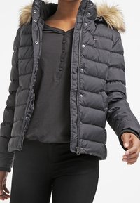 Černá puffová bunda s odnímatelnou umělou kožešinovou kapucí, zipovým zapínáním a horizontálním prošíváním. Nositá přes černý dlouhý rukáv.