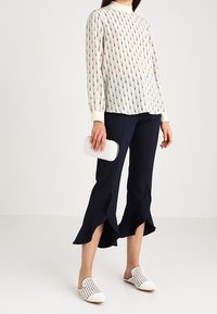 Ljus krämfärgad blus med vertikala marinblå tryck, marinblå utsvängda byxor, vita slip-on-skor och en strukturerad vit clutch-väska.