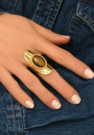 Bague en or adornée d'une pierre ovale en œil de tigre brun, avec des gravures détaillées et un large anneau, portée sur une main contre un fond en denim.