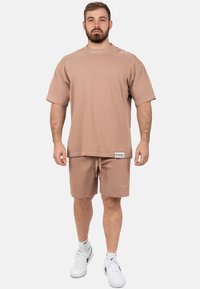 T-shirt oversized marrone chiaro con maniche corte e pantaloni abbinati. Materiale in cotone morbido e vita con cordino sui pantaloni. Branding su entrambi i pezzi.