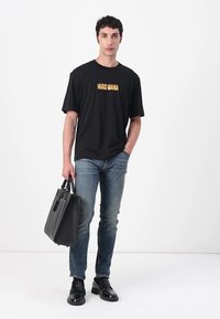 Svart T-skjorte med "HUGO MANIA" i gult, kombinert med blå jeans og svarte lærsko, mens han holder en svart bag. Enkelt og moderne design.