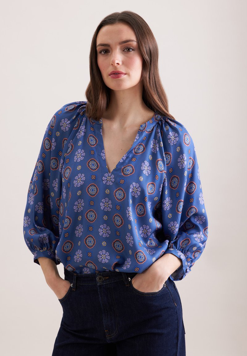 Femme aux longs cheveux bruns portant un chemisier bleu à motif floral et un jean foncé, les mains dans les poches, debout devant un fond uni.