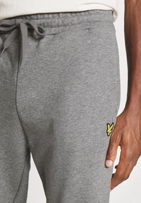 Närbild av en person som bär grå sweatpants med en knuten dragsko och en liten gul fågel-logotyp på höger lår.