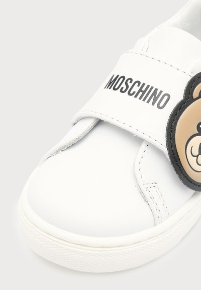 Baskets blanches Moschino avec une bride en Velcro avec un texte noir et un détail visage d'ours brun sur le côté.